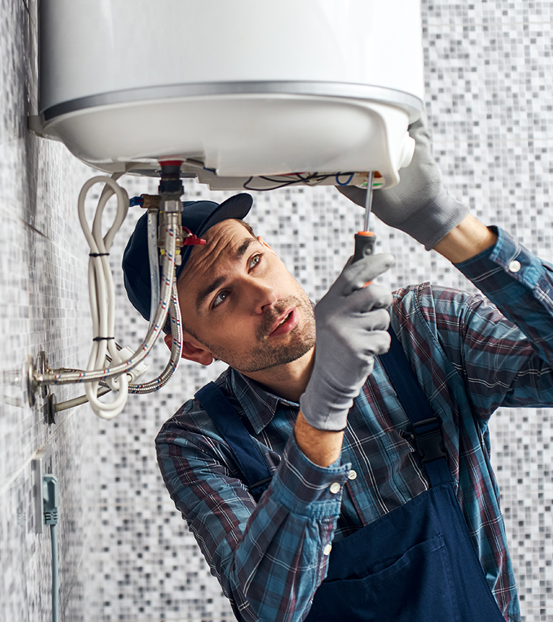 A technician in work gloves and a cap uses a screwdriver to repair or install a wall-mounted water heater in a tiled room.