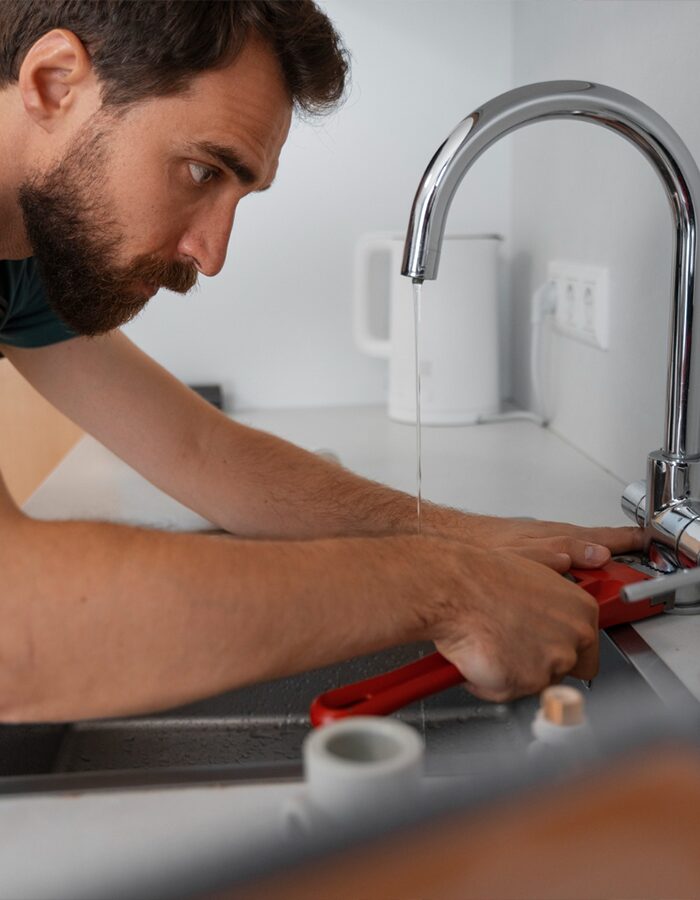 A man uses a red pipe wrench to plumber san francisco fix a kitchen sink faucet with running water.