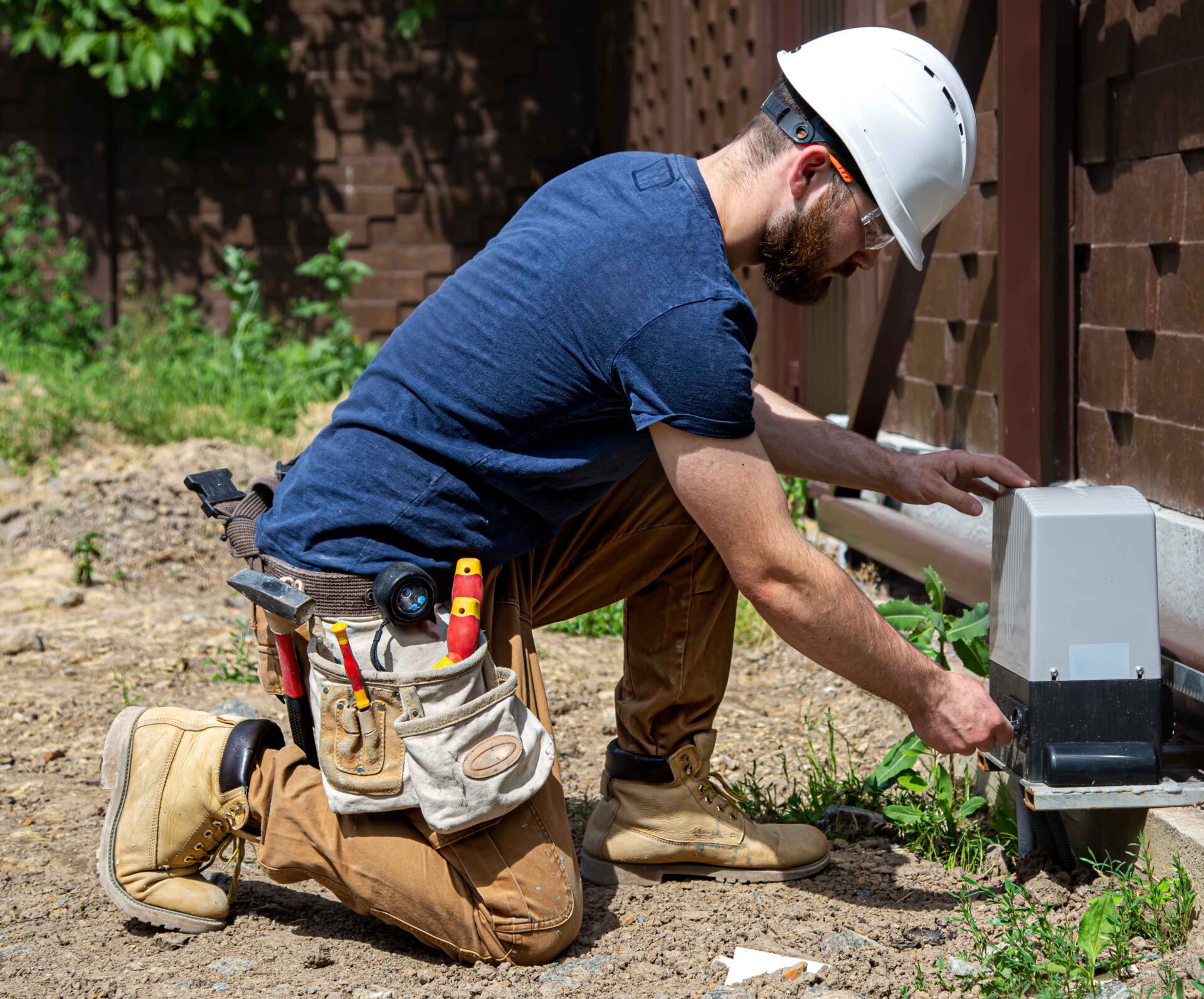 A sewer line repair san francisco construction worker wearing a hard hat and tool belt kneels on the ground while working on an outdoor electrical box near a fence.
