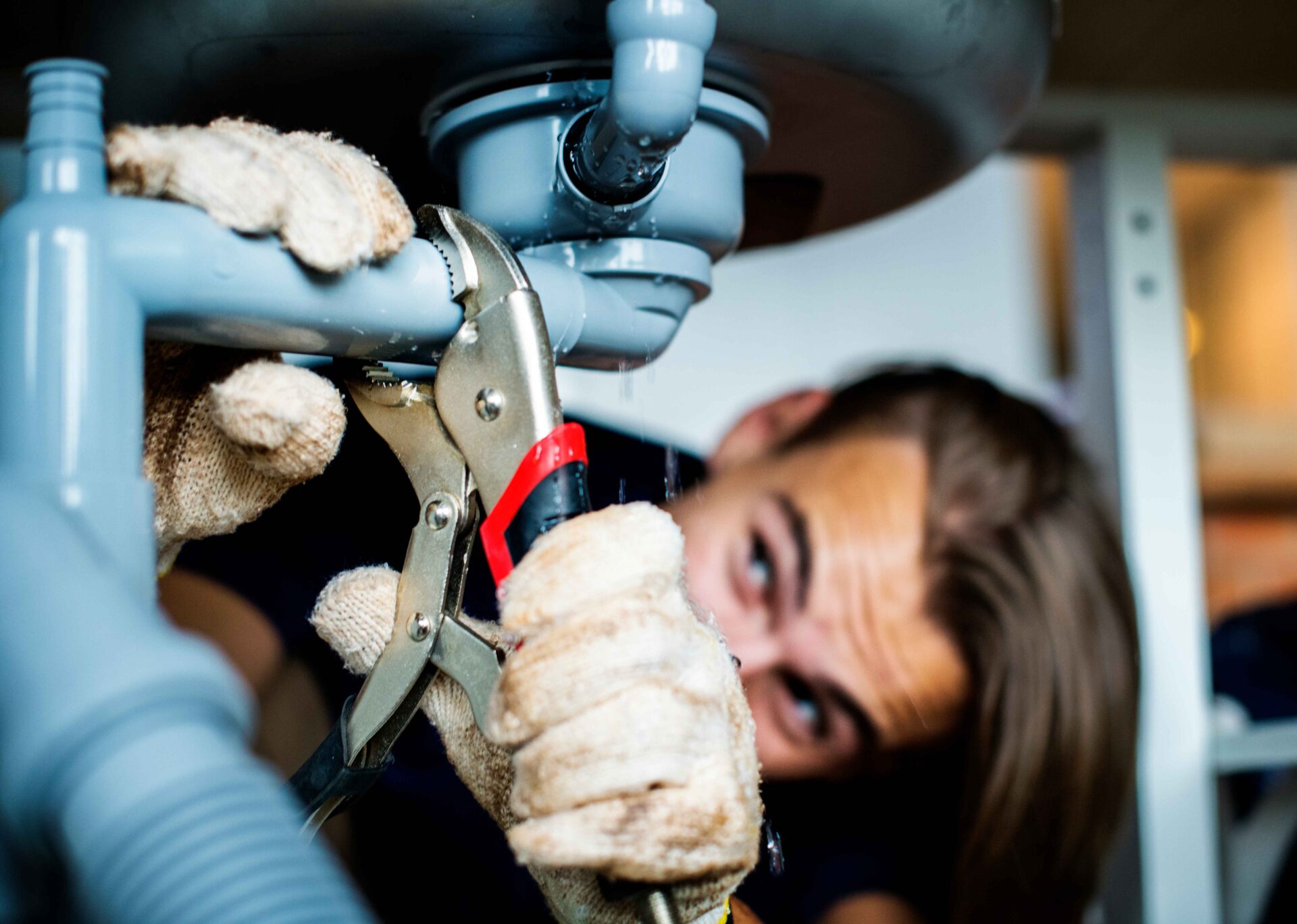 A person wearing gloves uses pliers to repair plumbing pipes under a sink plumber daly city.