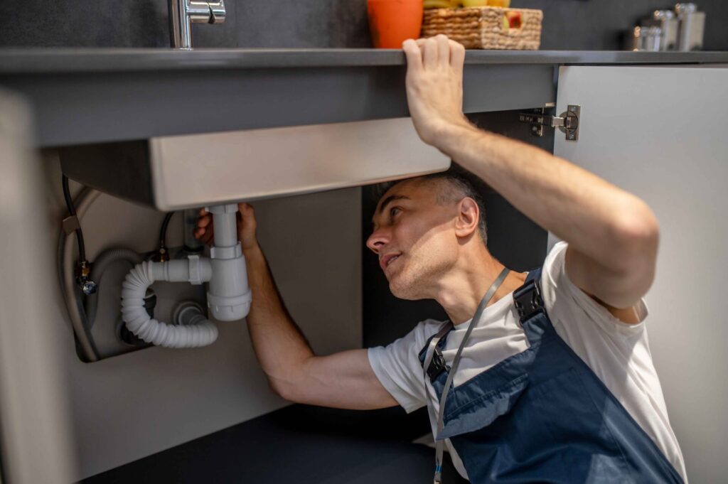 A drain cleaning san francisco man wearing overalls inspects plumbing under a kitchen sink with the cabinet doors open.